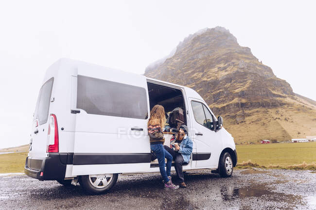 Хлопець і приваблива жінка в теплому одязі з кухлем, що дивиться далеко відкритими дверима фургона біля скелястого пагорба в Ісландії — стокове фото