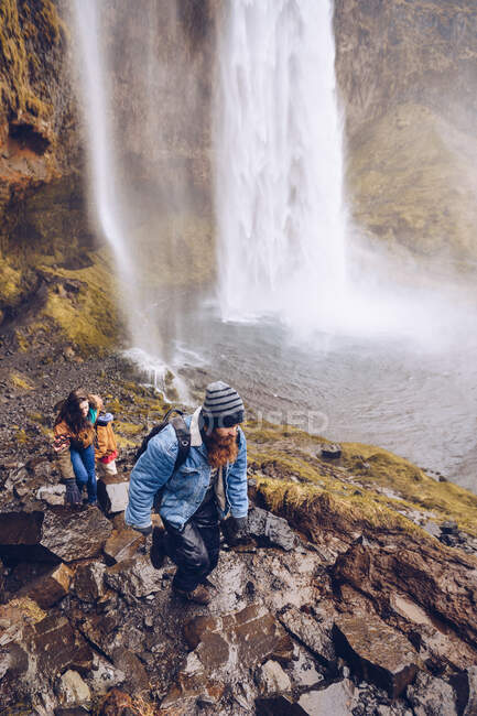 From above young lady and guy in warm wear scaling on hill near water cascade falling in river in Iceland — Stock Photo