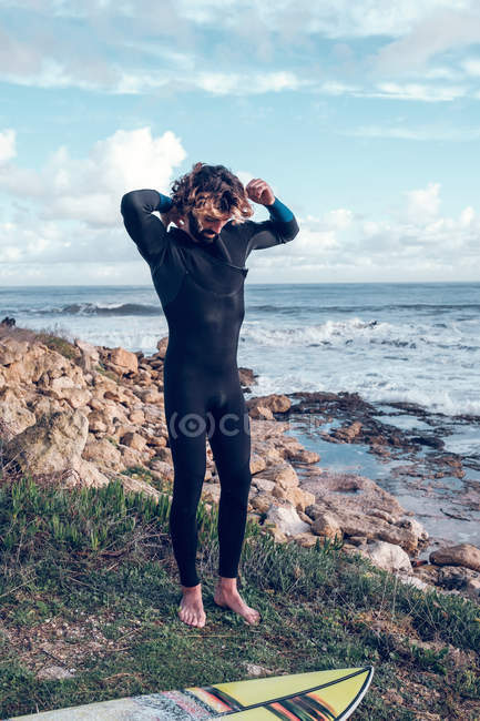 Young man putting on wetsuit on sea coast with surfboard — Stock Photo