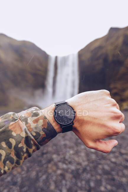 Mano recortada del tipo que muestra relojes negros cerca de la cascada de agua en Islandia sobre un fondo borroso - foto de stock