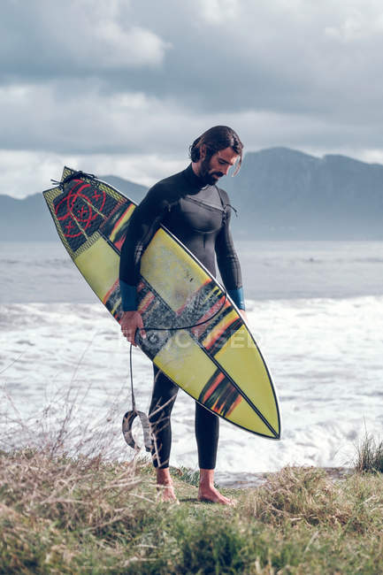 Joven en traje de neopreno con tabla de surf caminando por la costa del mar - foto de stock