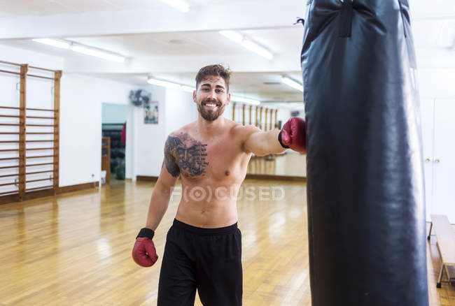 Retrato de un joven boxeador sonriente apoyado en un saco de boxeo en el gimnasio - foto de stock