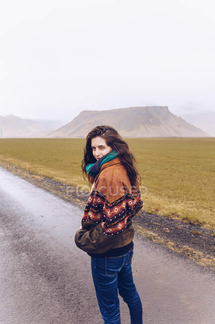 Vue de dos jeune femme attrayante regardant caméra sur la route entre les terres sauvages avec des collines de pierre en Islande — Photo de stock