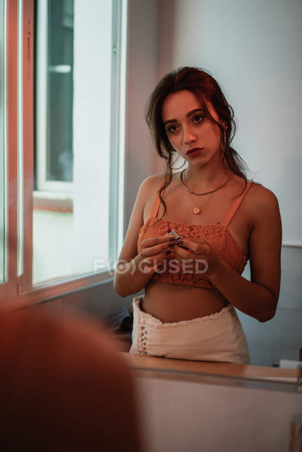 Charming young lady in tank top holding lipstick in front of mirror — Stock Photo