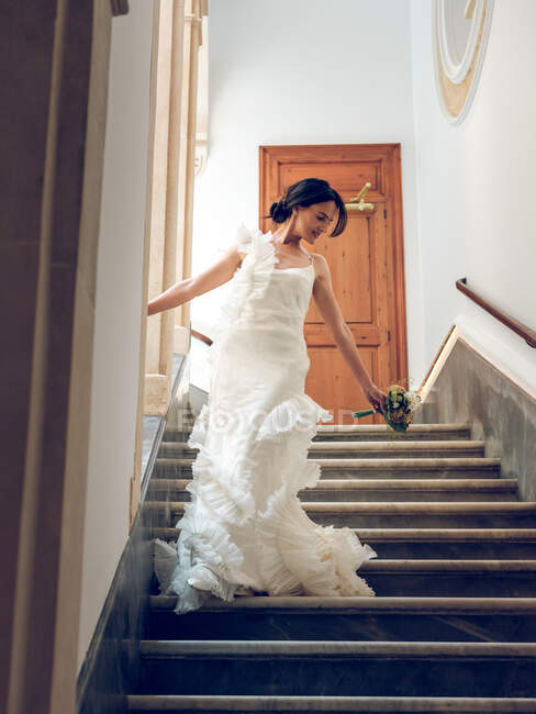 Hermosa mujer en vestido de novia elegante celebración de ramo y caminar por las escaleras del edificio de lujo - foto de stock