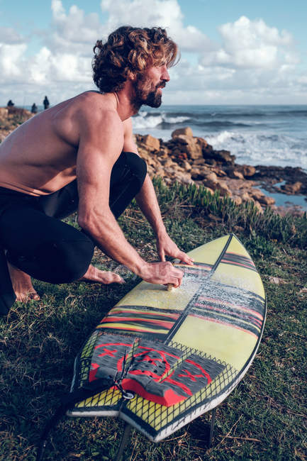 Junger Mann putzt Surfbrett an der Küste und blickt auf Aussicht — Stockfoto