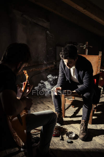 Парень, играющий на гитаре рядом с молодым человеком, курящим сигарету и использующим мобильный телефон на стуле на чердаке — стоковое фото