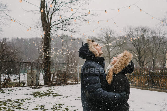 Side view of happy guy hugging young lady in ski jackets near wood decorated by illuminated fairy lights — Stock Photo