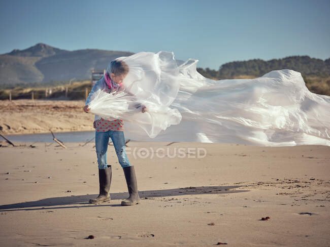 Concepto de niño enredado en textil blanco ondeando por el viento en la costa de arena sobre el fondo de montaña - foto de stock
