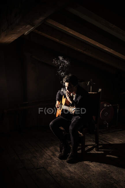 Chico tocando en la guitarra y fumar cigarrillo en la silla en el desván en la oscuridad - foto de stock