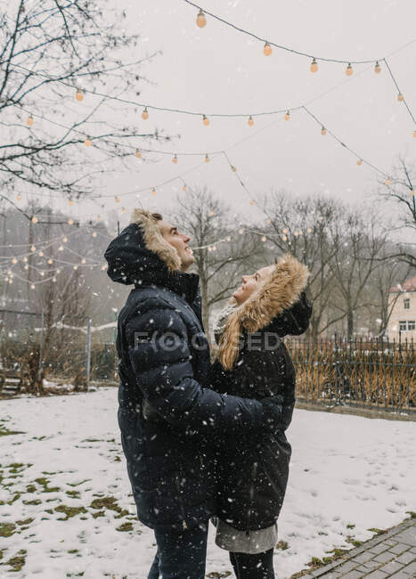 Vue latérale du mec heureux embrassant jeune dame dans des vestes de ski près du bois décoré par des lumières de fées éclairées — Photo de stock