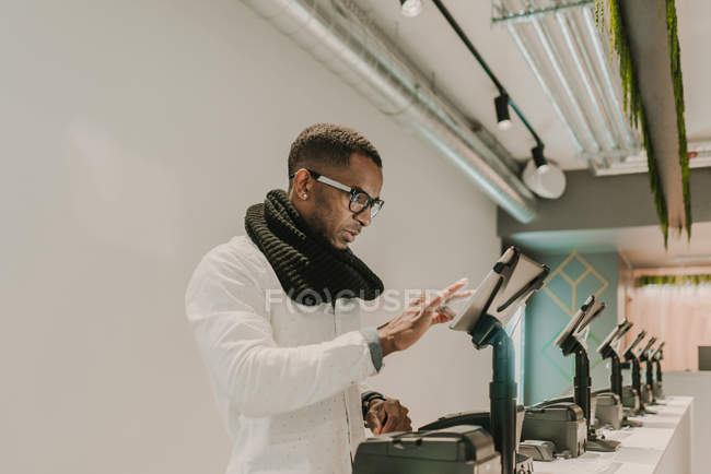 Side view of handsome African American man in trendy outfit standing near counter in stylish shop and using cash register — Stock Photo