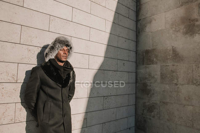 Stylish African American man in fur hat leaning on brick wall on sunny day — Stock Photo