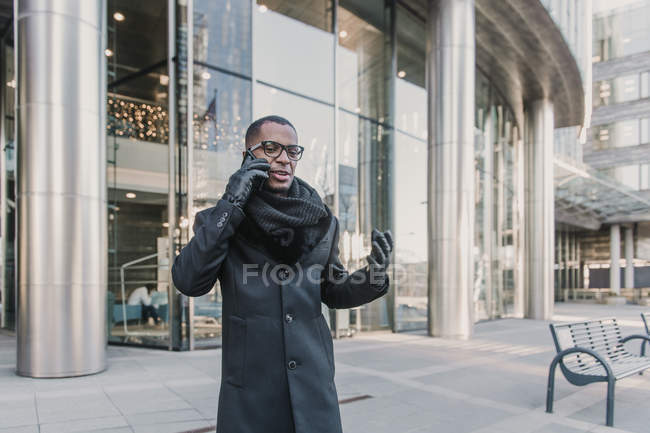 Handsome African American businessman in stylish warm clothes talking on smartphone  on street of modern city — Stock Photo