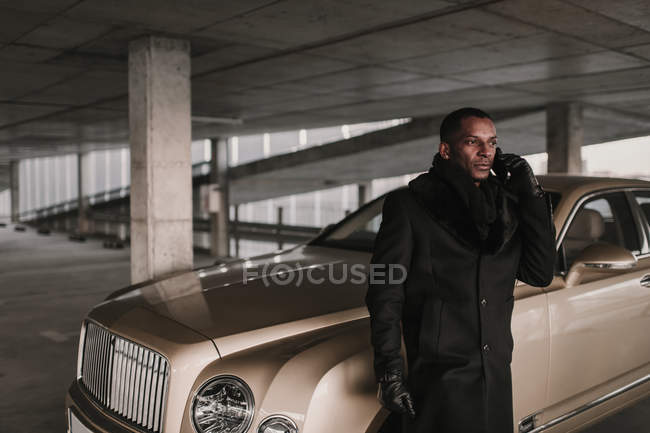 Confiado hombre de negocios afroamericano con un abrigo elegante de pie cerca del vehículo de lujo en el estacionamiento y hablando en el teléfono inteligente - foto de stock