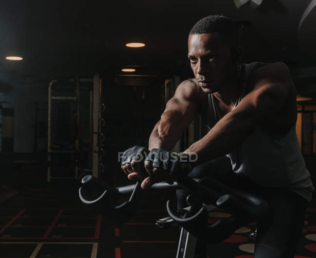 Confident African American man sitting on exercise bike during workout in dark gym — Stock Photo
