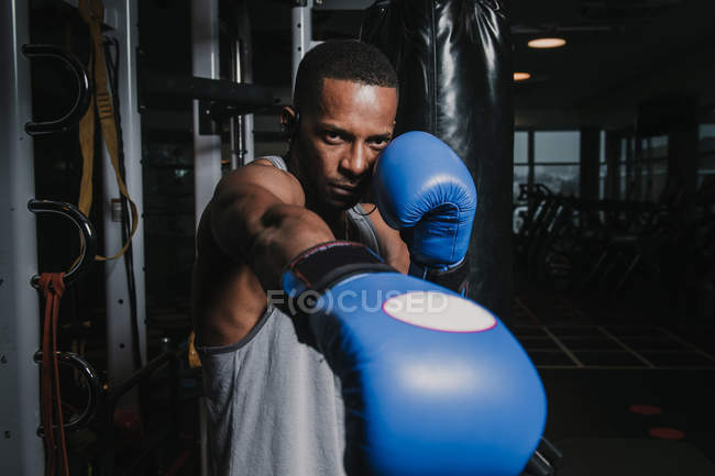 Uomo nero in blu guantoni da boxe punzonatura aria mentre si allena in palestra moderna scura — Foto stock