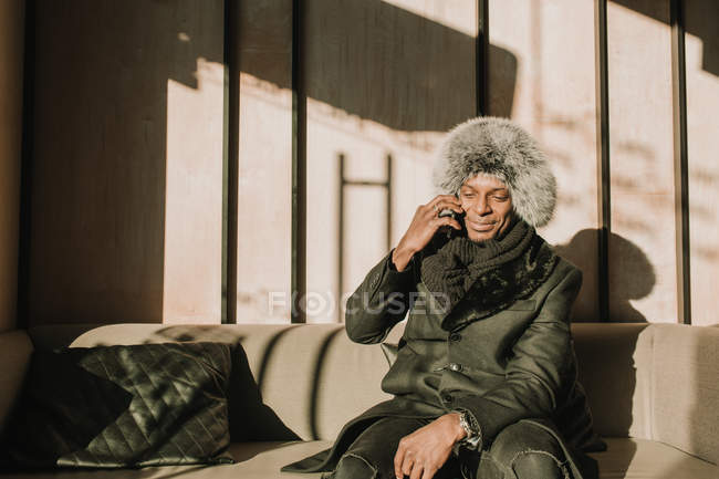 Красивый афроамериканец в теплой одежде держит чашку горячего напитка и смотрит в сторону, сидя на удобном диване в кафе — стоковое фото