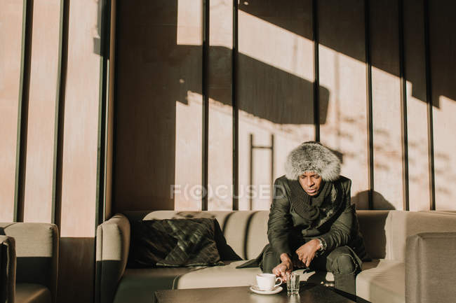 Stylish African American male in warm clothes sitting on comfortable sofa in cafe — Stock Photo