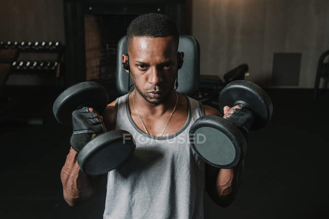 Junger afrikanisch-amerikanischer Mann hebt zwei schwere Hanteln und hört während des Trainings in einem modernen Fitnessstudio Musik — Stockfoto