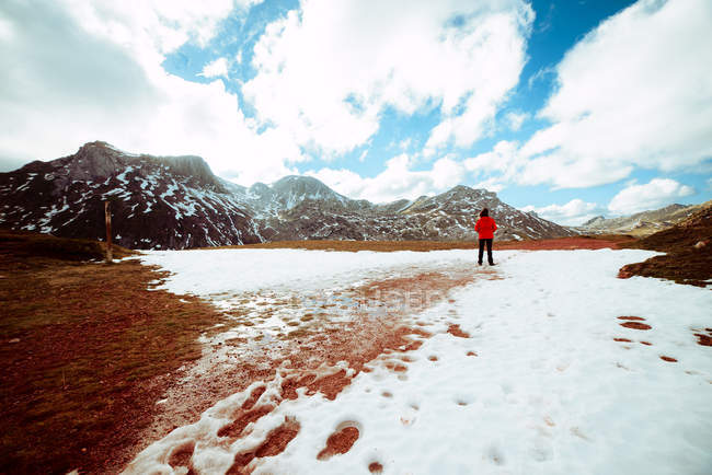 Back view of female in red jacket standing on melting snow against beautiful cloudy sky and amazing mountain ridge — Stock Photo