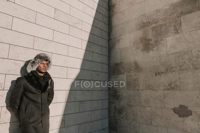 African American man in fur hat leaning on brick wall on sunny day on city street — Stock Photo