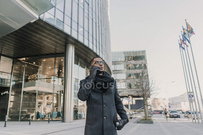 Hombre afroamericano guapo en ropa de abrigo elegante gestos y hablar en el teléfono inteligente, mientras que de pie en la calle de la ciudad moderna - foto de stock
