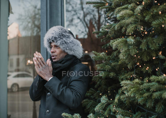 Schöner afrikanisch-amerikanischer Mann in warmer Kleidung reibt sich die Hände, während er in der Nähe des Weihnachtsbaums mit Lichterketten auf der Stadtstraße steht — Stockfoto