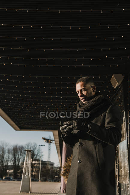 From below shot of handsome African American man using modern smartphone while standing near modern building decorating for Christmas with fairy light garlands — Stock Photo