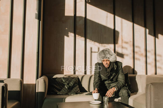 Hombre afroamericano guapo en traje cálido sosteniendo una taza de bebida caliente y mirando hacia otro lado mientras está sentado en un cómodo sofá en la cafetería - foto de stock