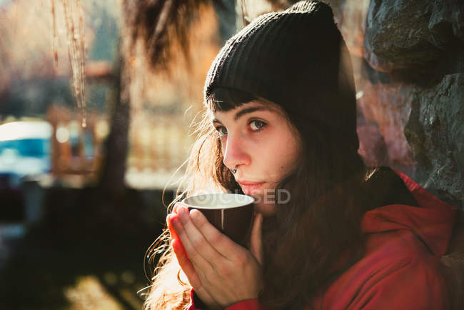 Вид сбоку на красивую молодую женщину в стильной шляпе, пахнущую ароматным горячим напитком, сидя в сельской местности в солнечный день — стоковое фото