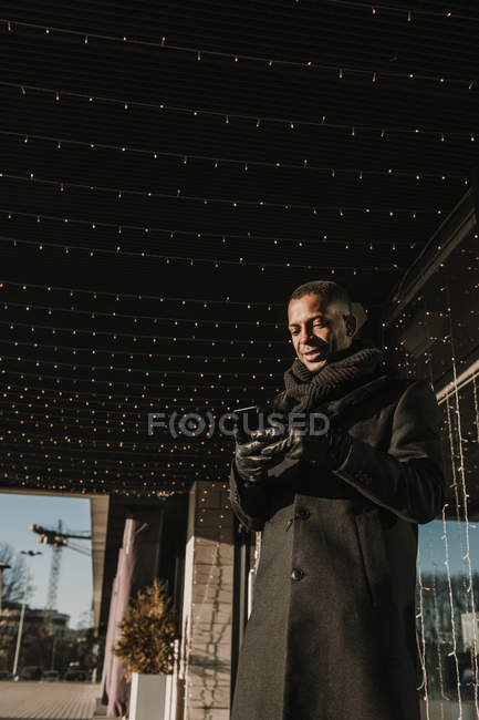 From below shot of handsome African American man using modern smartphone while standing near modern building — Stock Photo