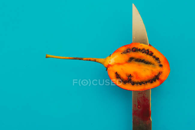 Sharp knife slicing tasty ripe tamarillo on bright blue background — Stock Photo