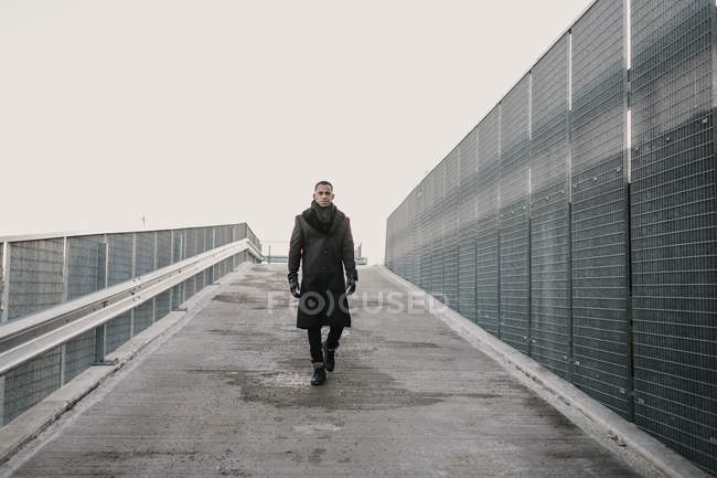 African American man in trendy clothes walking on wet asphalt near fence on gray day in city — Stock Photo