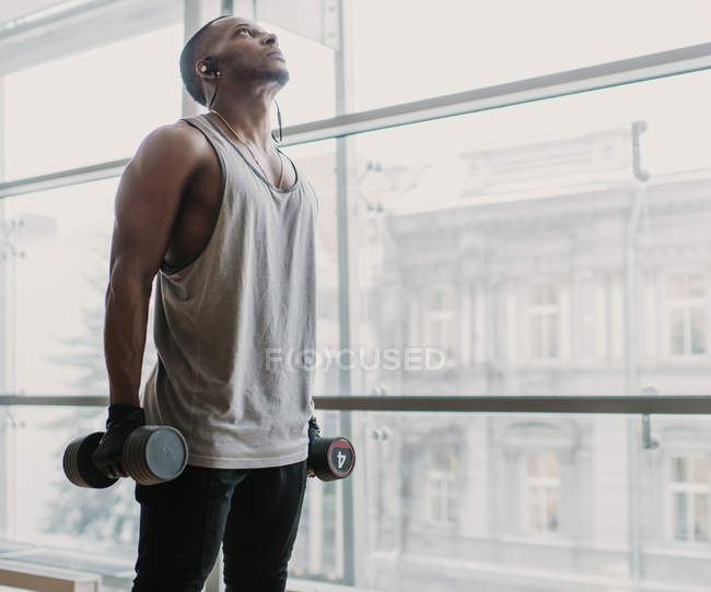 Young African American male with two dumbbells listening to music and looking up while standing near window in modern gym — Stock Photo