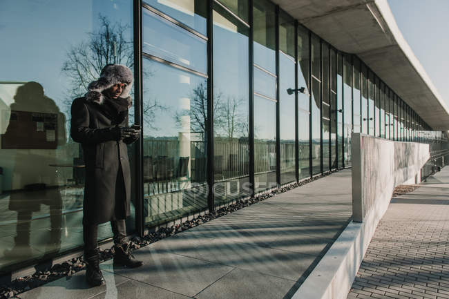 Black man in warm clothes standing near glass wall of modern building and browsing smartphone on sunny day on city street — Stock Photo