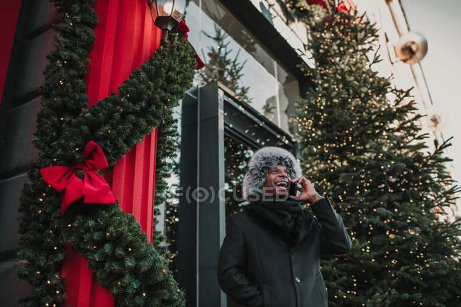 Afrikanisch-amerikanischer Mann in warmer Kleidung spricht auf Smartphone, während er auf der Stadtstraße in der Nähe eines Gebäudes steht und einen weihnachtlich geschmückten Nadelbaum hat — Stockfoto