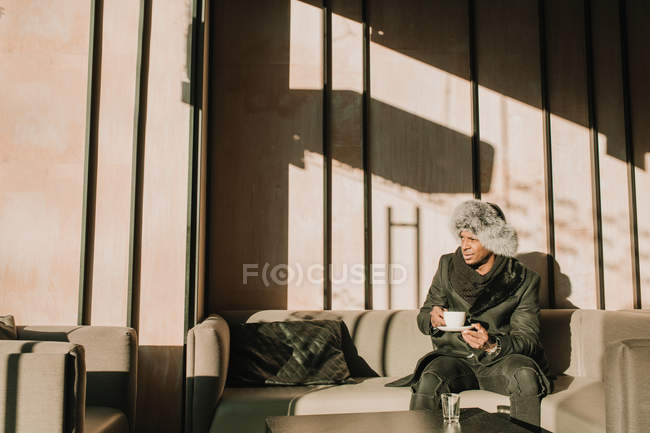 Bonito homem afro-americano em roupa quente segurando xícara de bebida quente e olhando para longe enquanto sentado no sofá confortável no café — Fotografia de Stock