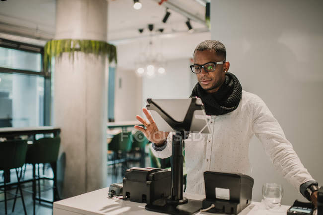 Handsome African American man in trendy outfit standing near counter in stylish shop and using cash register — Stock Photo