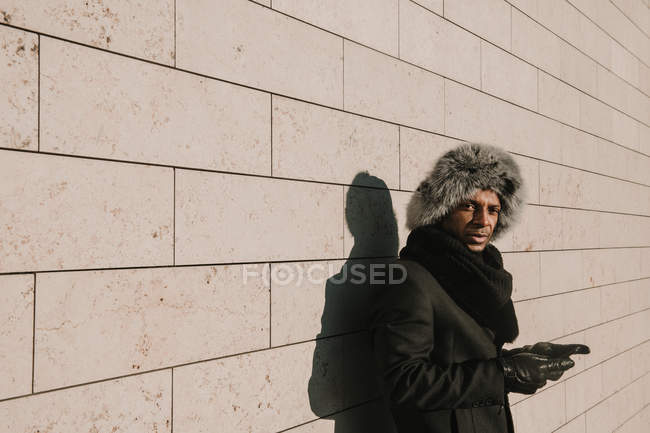 Attractive African American man in fur hat leaning on brick wall with mobile phone on sunny day on city street — Stock Photo