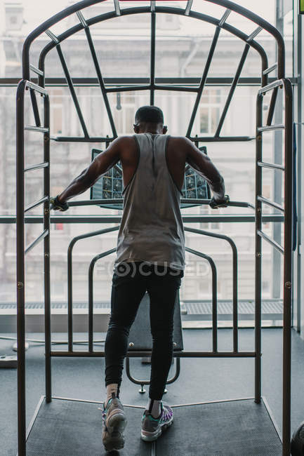 Vista trasera del chico afroamericano en ropa deportiva usando equipo de gimnasio mientras hace ejercicio cerca de la ventana - foto de stock