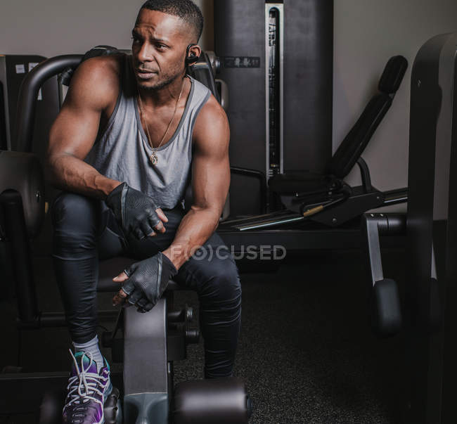 Atraente cara afro-americano em sportswear ouvir música em fones de ouvido e olhando para longe no ginásio moderno — Fotografia de Stock