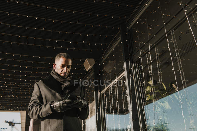 Afro-Américain utilisant un smartphone moderne tout en se tenant près de bâtiment moderne décoration pour Noël avec des guirlandes de lumière de fée — Photo de stock
