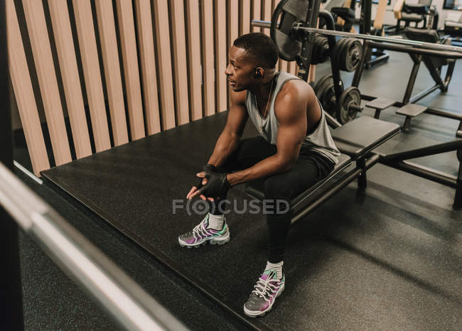 Homem preto em fones de ouvido sentado na máquina de exercício — Fotografia de Stock