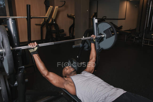 Schöner afrikanisch-amerikanischer Mann in Sportbekleidung, der auf einer Bank liegt und schwere Langhantel hebt, während er im modernen Fitnessstudio trainiert — Stockfoto