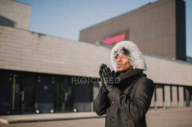 Привабливий афроамериканець у хутряному капелюсі стоїть біля сучасного будинку в сонячний день на вулицях міста, потираючи руки. — стокове фото