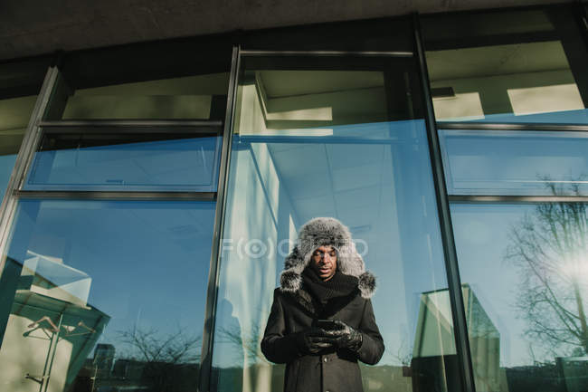 Uomo nero in abiti caldi in piedi vicino alla parete di vetro di edificio moderno e smartphone di navigazione nella giornata di sole sulla strada della città — Foto stock