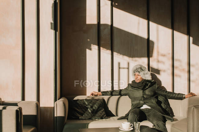 Hombre afroamericano guapo en traje cálido mirando hacia otro lado mientras está sentado en un cómodo sofá en la cafetería - foto de stock