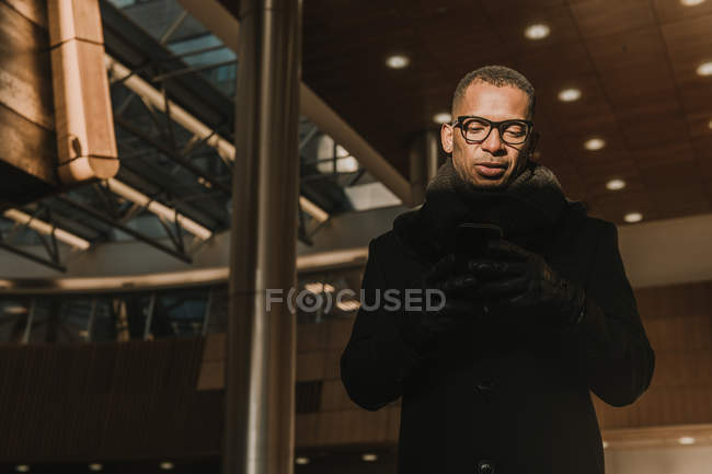 D'en bas plan de bel homme afro-américain en utilisant un téléphone portable debout près du bâtiment moderne — Photo de stock