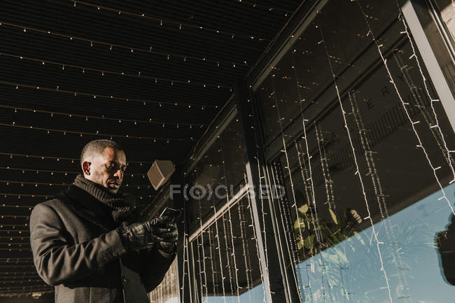 African American man using modern smartphone while standing near modern building decorating for Christmas with fairy light garlands — Stock Photo